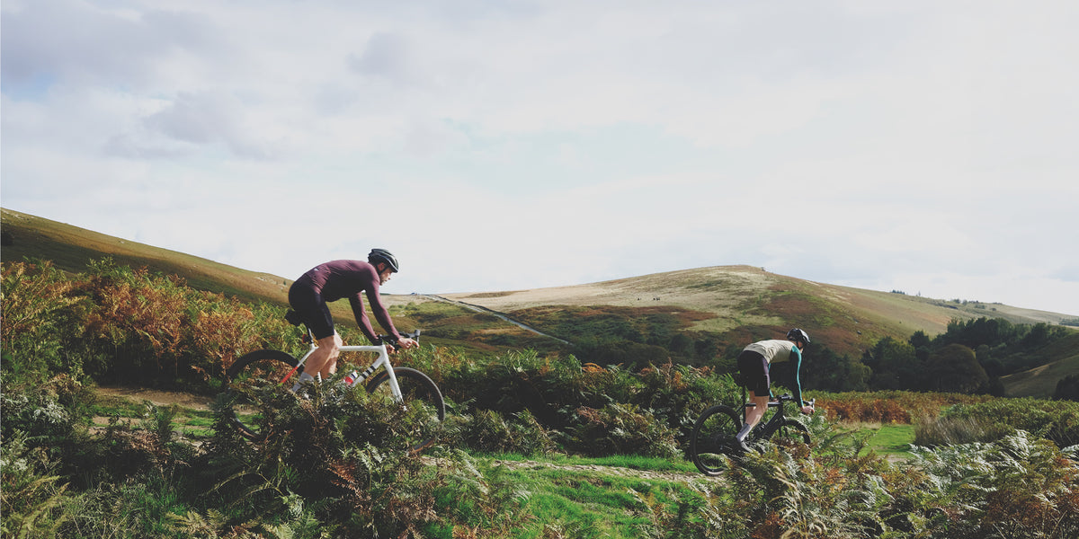 Two cyclists ride a grassy track through autumn bracken across rolling moorland hills under a wide, cloudy sky